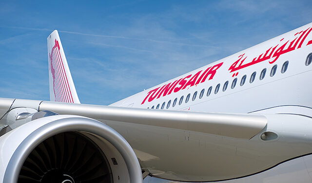 Photo d'un avion Tunisair au sol, de préférence sur le tarmac de l'aéroport de Tunis-Carthage. Prise en lumière naturelle, livrée blanche et rouge visible, logo de la compagnie bien lisible sur le fuselage ou la queue de l'appareil. Ambiance sobre, pas de passagers au premier plan.